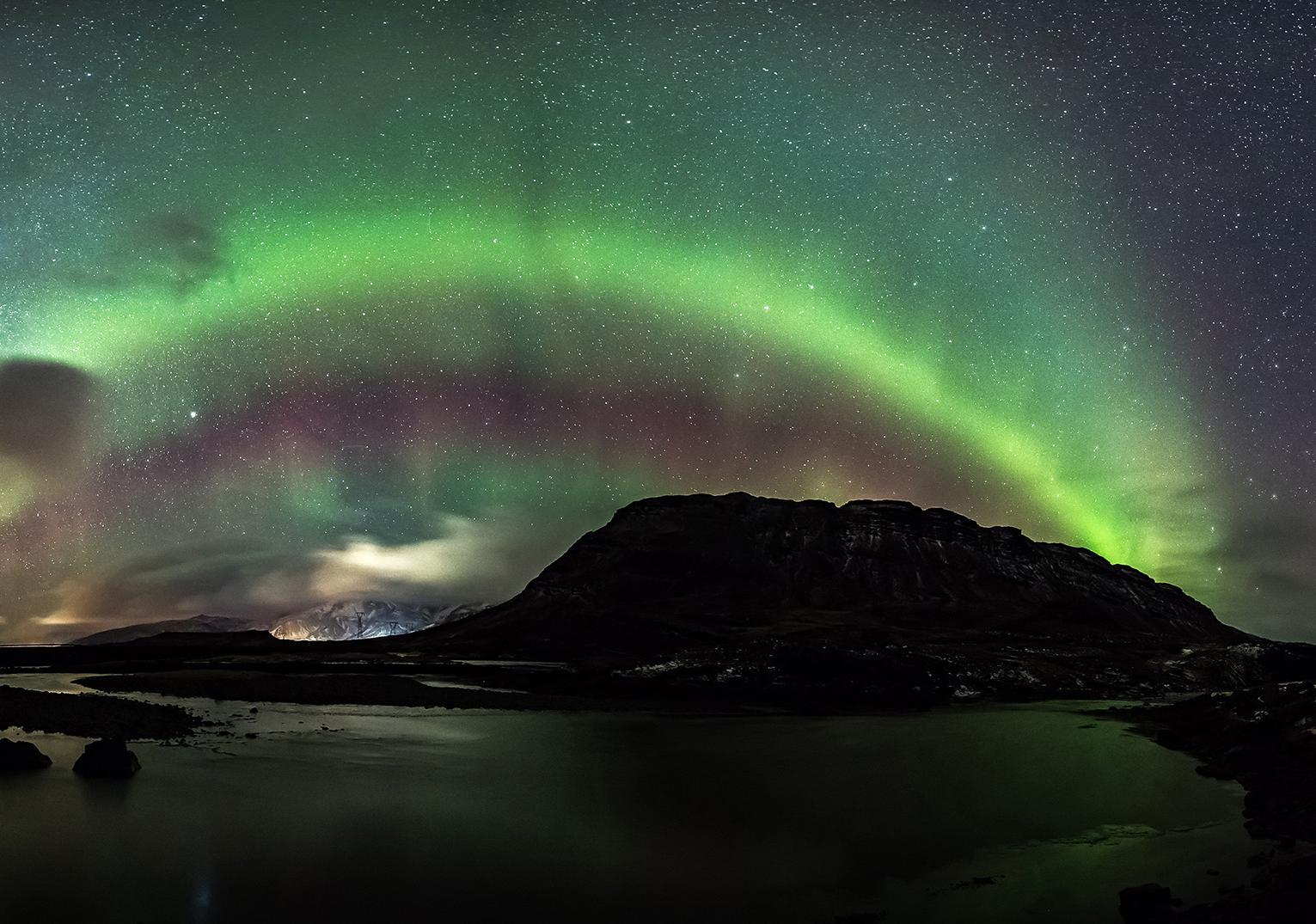 Large rocky cliff face with waterfall under a night sky glowing with green lights from the aurora borealis