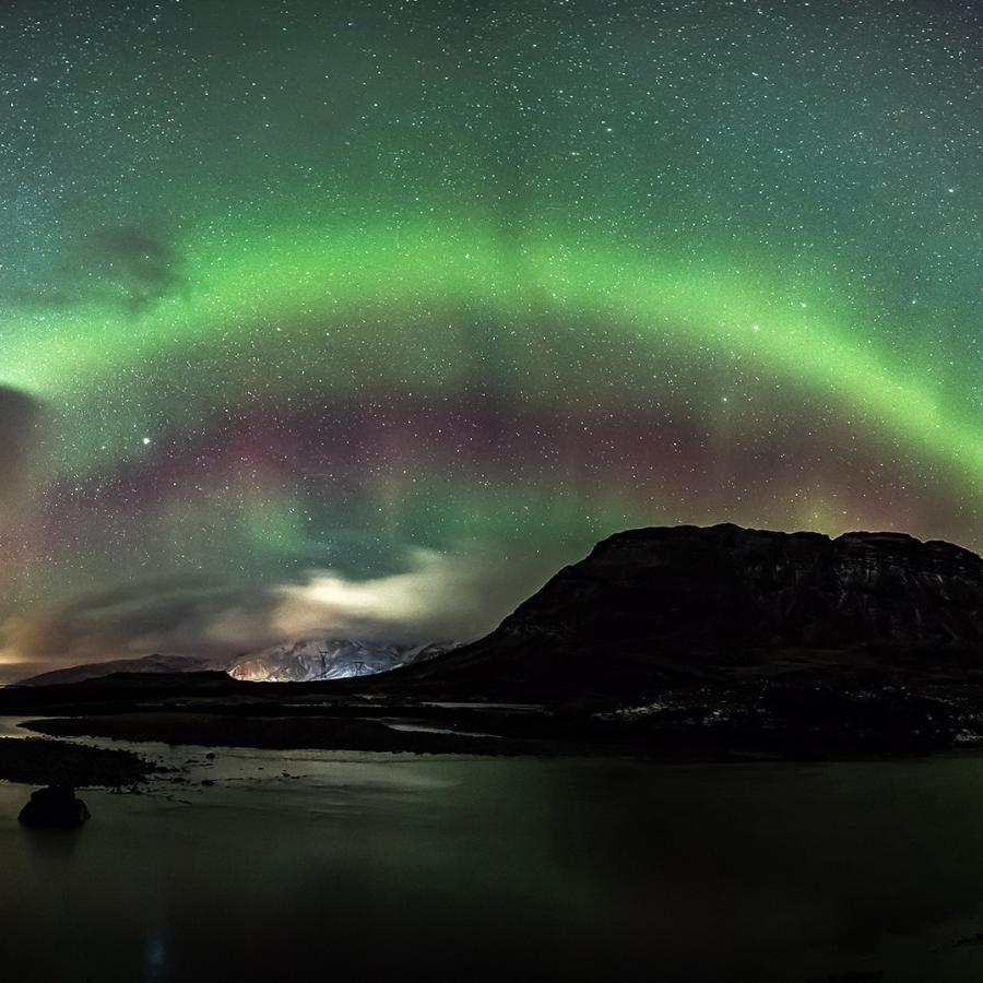Large rocky cliff face with waterfall under a night sky glowing with green lights from the aurora borealis