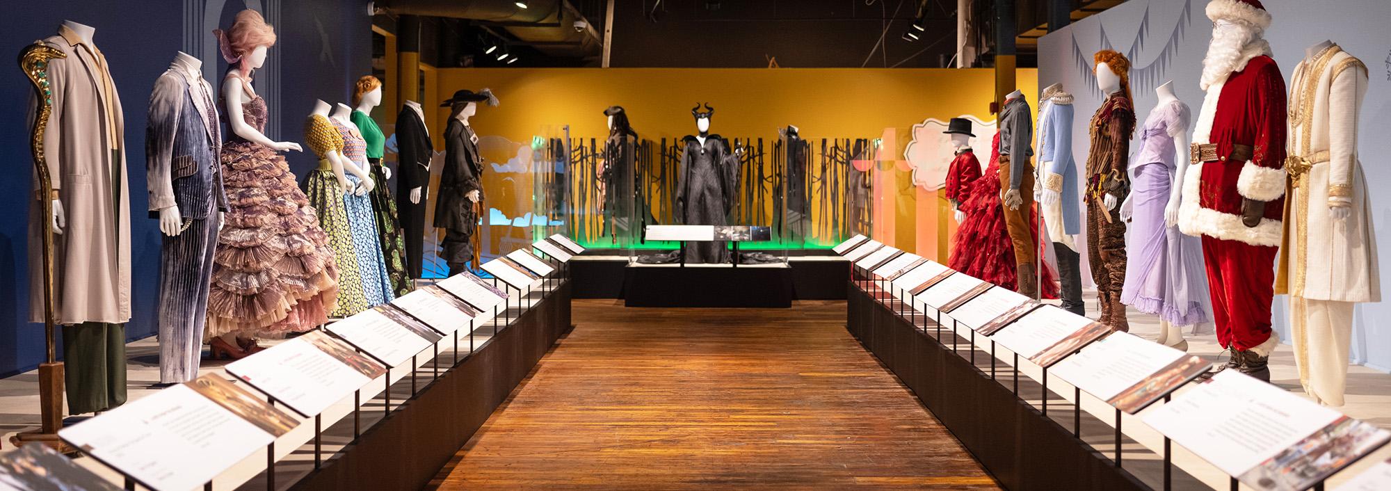 Aisle with wooden floor and a row of mannequins on raised platforms on either side. They are dressed in various costumes including a red and white Santa suit, a frilly lavender gown, a white and gold Aladdin suit.