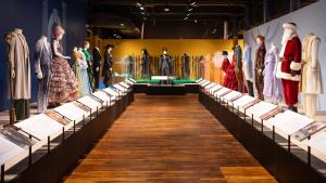 Aisle with wooden floor and a row of mannequins on raised platforms on either side. They are dressed in various costumes including a red and white Santa suit, a frilly lavender gown, a white and gold Aladdin suit.
