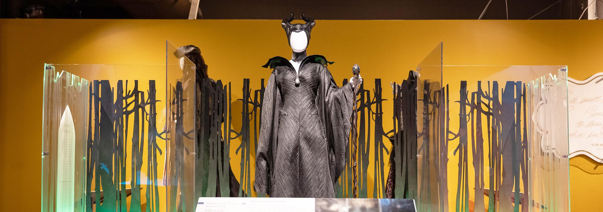 Platform with low panel in front. On the platform is a tall mannequin wearing a long and black gown with a large collar and a headdress featuring two curvy horns