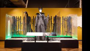 Platform with low panel in front. On the platform is a tall mannequin wearing a long and black gown with a large collar and a headdress featuring two curvy horns