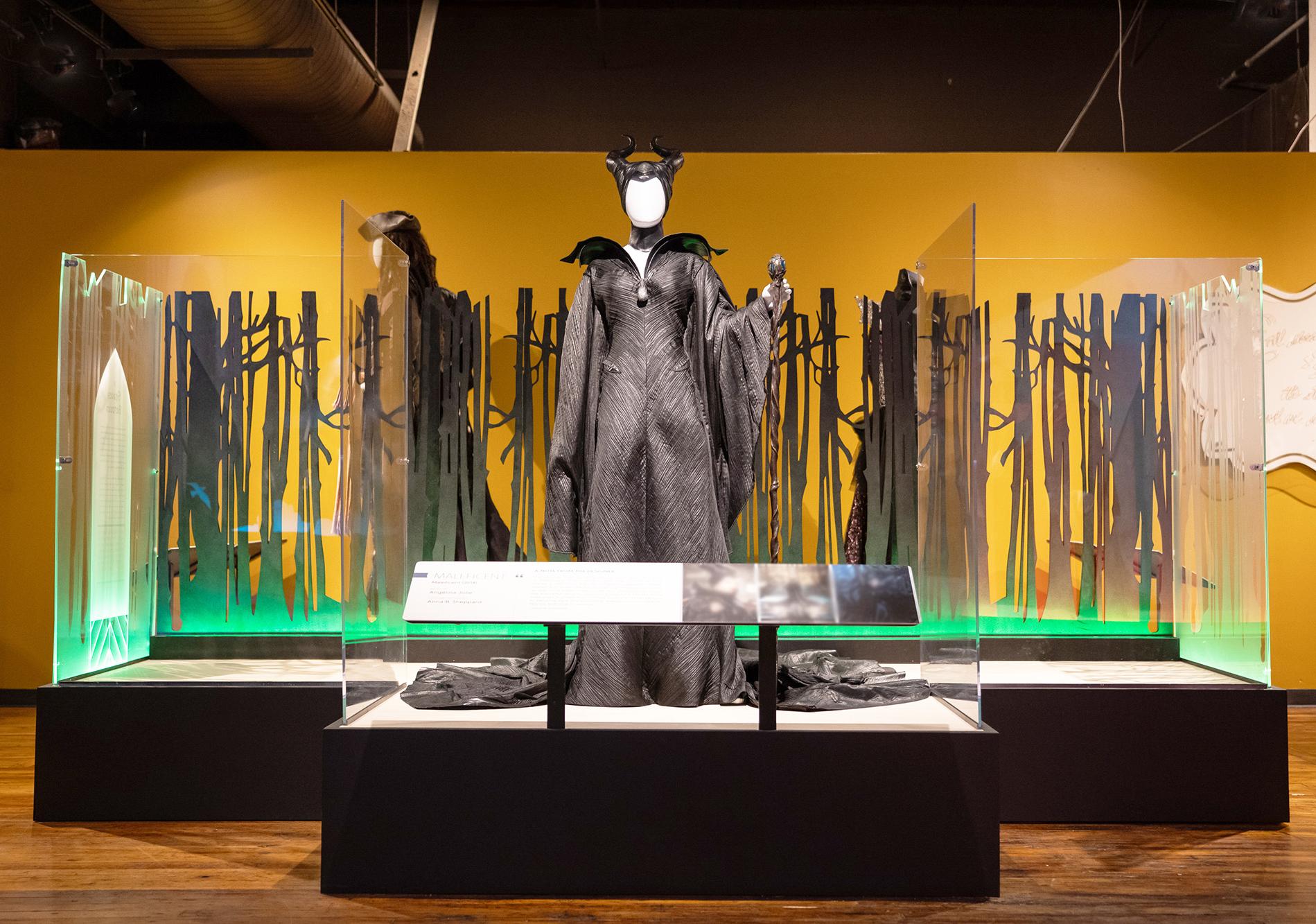 A man and woman stand looking up at a row of 7 mannequins that are dressed in various costumes, including a frilly lavender gown, a gray velvet suit, black and red robes with an embroidered cape.