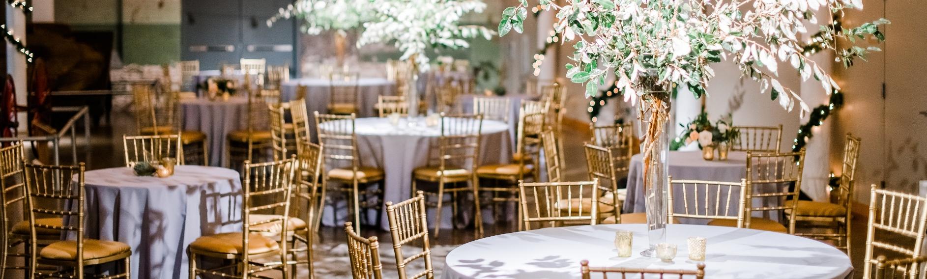 Large space with string lights suspended from ceiling and round tables with silver table cloths, gold chairs and large flower center pieces.