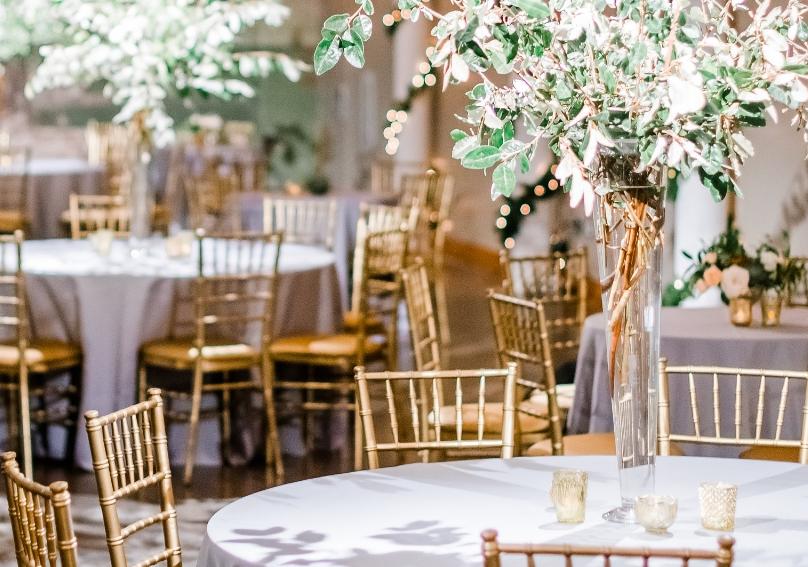 Large space with string lights suspended from ceiling and round tables with silver table cloths, gold chairs and large flower center pieces.