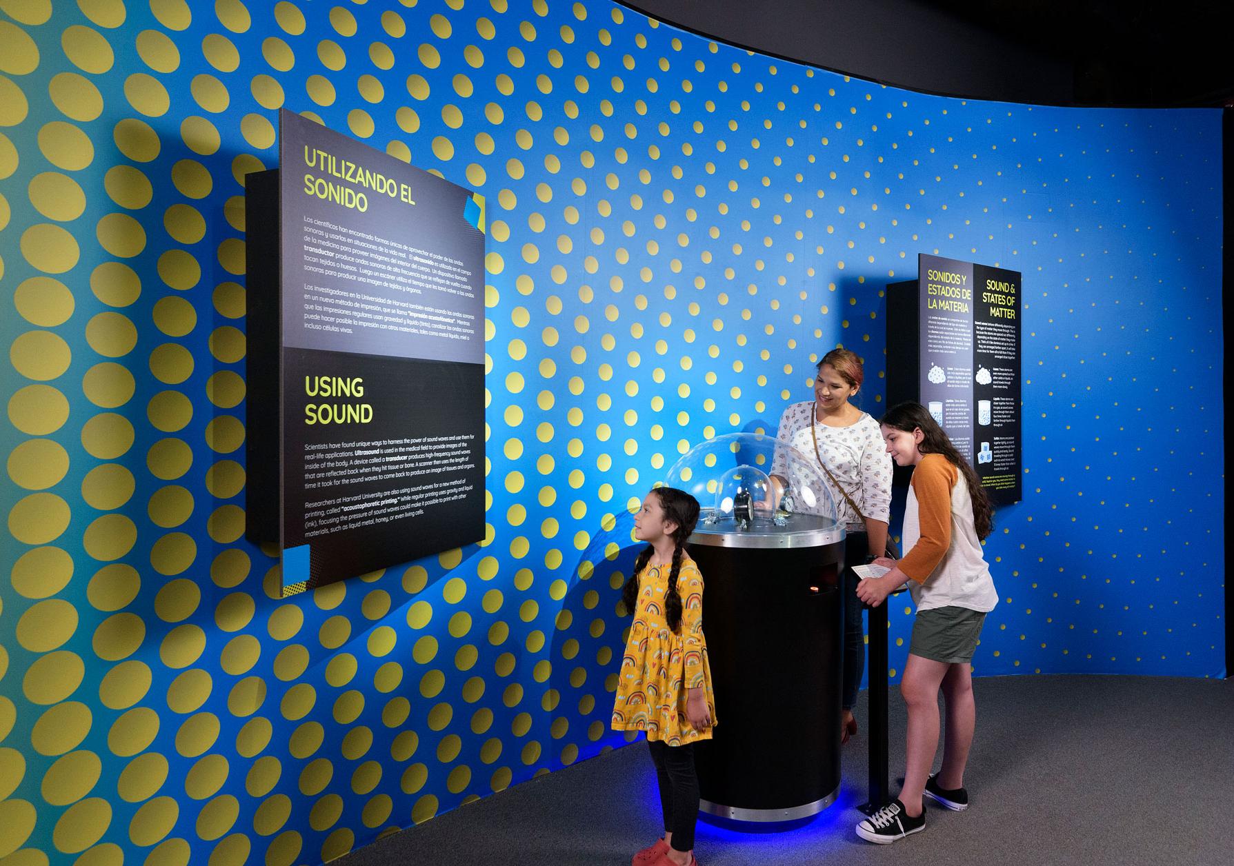 A mother and her two daughters look at hands-on demonstration and read a wall text panel in the Science of Sound exhibit.