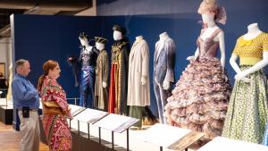 A man and woman stand looking up at a row of 7 mannequins that are dressed in various costumes, including a frilly lavender gown, a gray velvet suit, black and red robes with an embroidered cape.
