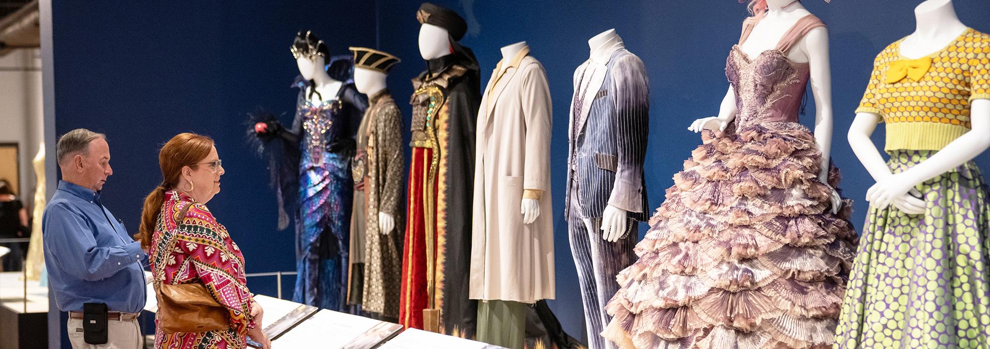 A man and woman stand looking up at a row of 7 mannequins that are dressed in various costumes, including a frilly lavender gown, a gray velvet suit, black and red robes with an embroidered cape.