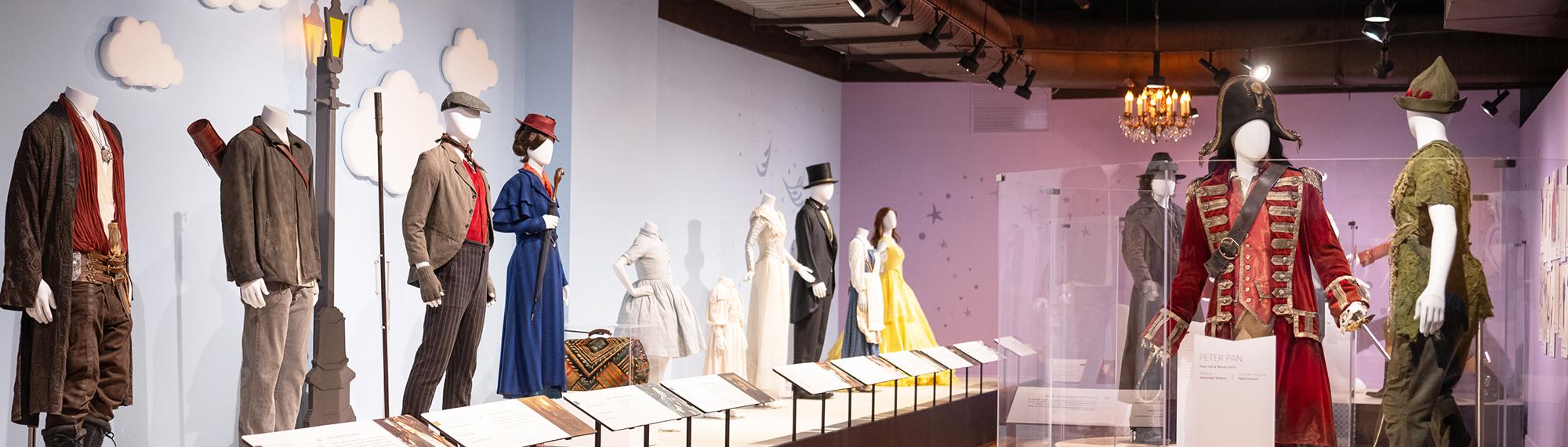 Side view of mannequin wearing voluminous pale blue ball gown with small butterfly appliques across the bodice. Two women stand on the right, one in a blue and white dress and the other in black pants and patterned blouse, stand looking at the ball gown.