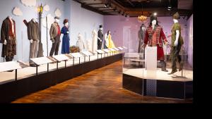 Room with hard wooden flooring and a row of platforms displaying 8 mannequins in various costumes, including weathered pants and brown jacket, a royal blue women's dress suit with red heeled shoes, a white slim feathered gown and more. Two mannequins on a platform to the rights side are wearing a pirate costume with a red velvet embroidered coat and bicorn hat and the other figure (Peter Pan) has forest green trousers boots, and a hat.