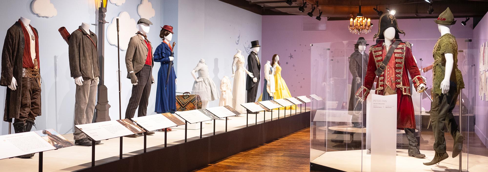 Room with hard wooden flooring and a row of platforms displaying 8 mannequins in various costumes, including weathered pants and brown jacket, a royal blue women's dress suit with red heeled shoes, a white slim feathered gown and more. Two mannequins on a platform to the rights side are wearing a pirate costume with a red velvet embroidered coat and bicorn hat and the other figure (Peter Pan) has forest green trousers boots, and a hat. 
