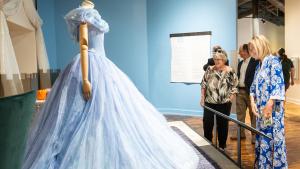 Side view of mannequin wearing voluminous pale blue ball gown with small butterfly appliques across the bodice. Two women stand on the right, one in a blue and white dress and the other in black pants and patterned blouse, stand looking at the ball gown.