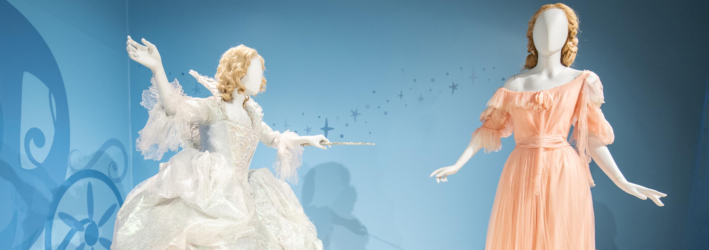 Two mannequins stand with one dressed in a voluminous light silvery white gown holding a thin wand pointed at the other figure which is wearing a tattered pale pink dress. 