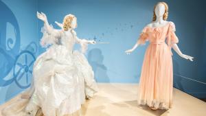 Two mannequins stand with one dressed in a voluminous light silvery white gown holding a thin wand pointed at the other figure which is wearing a tattered pale pink dress.