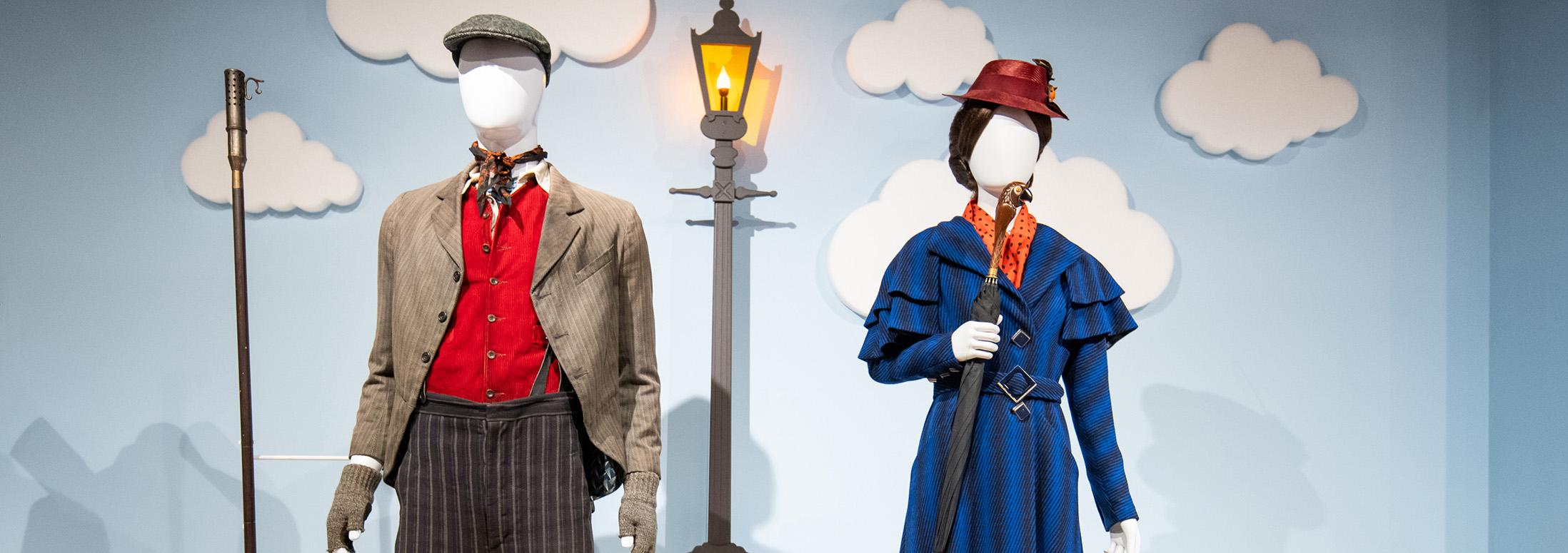 Two mannequins on plaform. The wall behind them in plae blue and decorate with clouds and the image of a light post lit with a gas flame. One figure is dress in brown striped trousers, a red vest and a tan dirty jacket. The other is dressed in a royal blue ladies' dress suit with red heeled shoes and a red hat. She is holding an umbrella with a handle carved like bird's head.