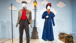Two mannequins on plaform. The wall behind them in plae blue and decorate with clouds and the image of a light post lit with a gas flame. One figure is dress in brown striped trousers, a red vest and a tan dirty jacket. The other is dressed in a royal blue ladies' dress suit with red heeled shoes and a red hat. She is holding an umbrella with a handle carved like bird's head.