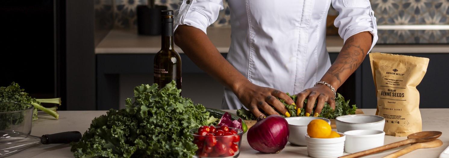 Woman in white chef jacket is handling greens with a table of cooking dishes in front of her