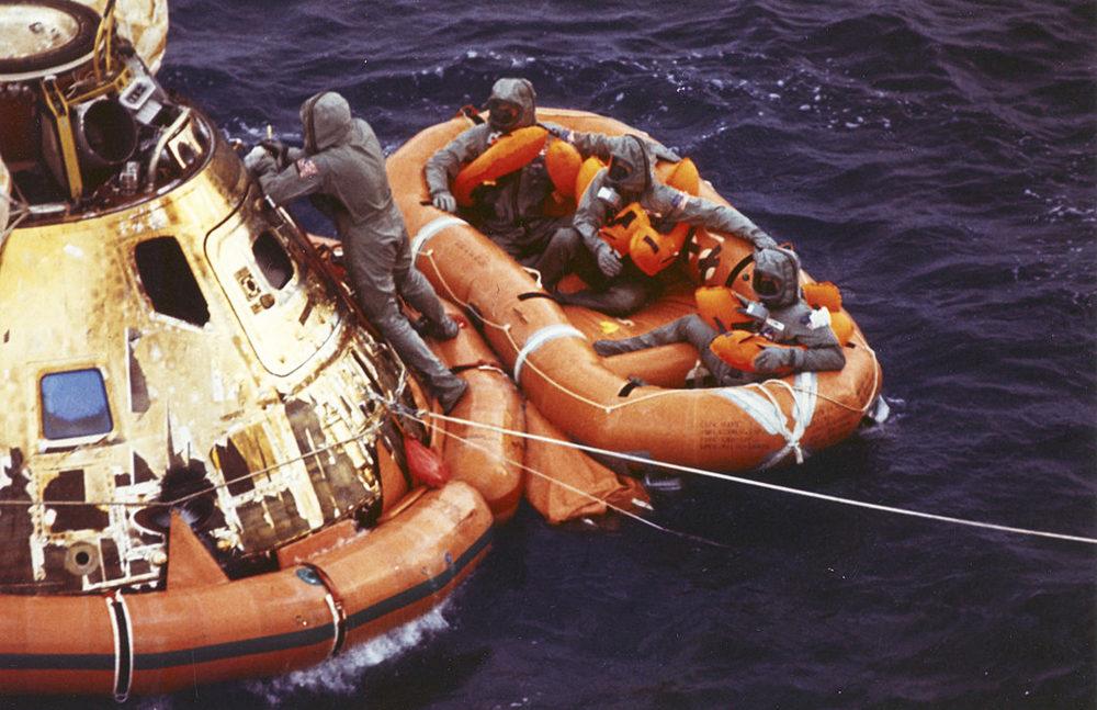 Space capsule and rescue raft shown in water. One individual is still on the capsule, three are in the raft.