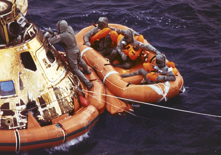 Space capsule and rescue raft shown in water. One individual is still on the capsule, three are in the raft.