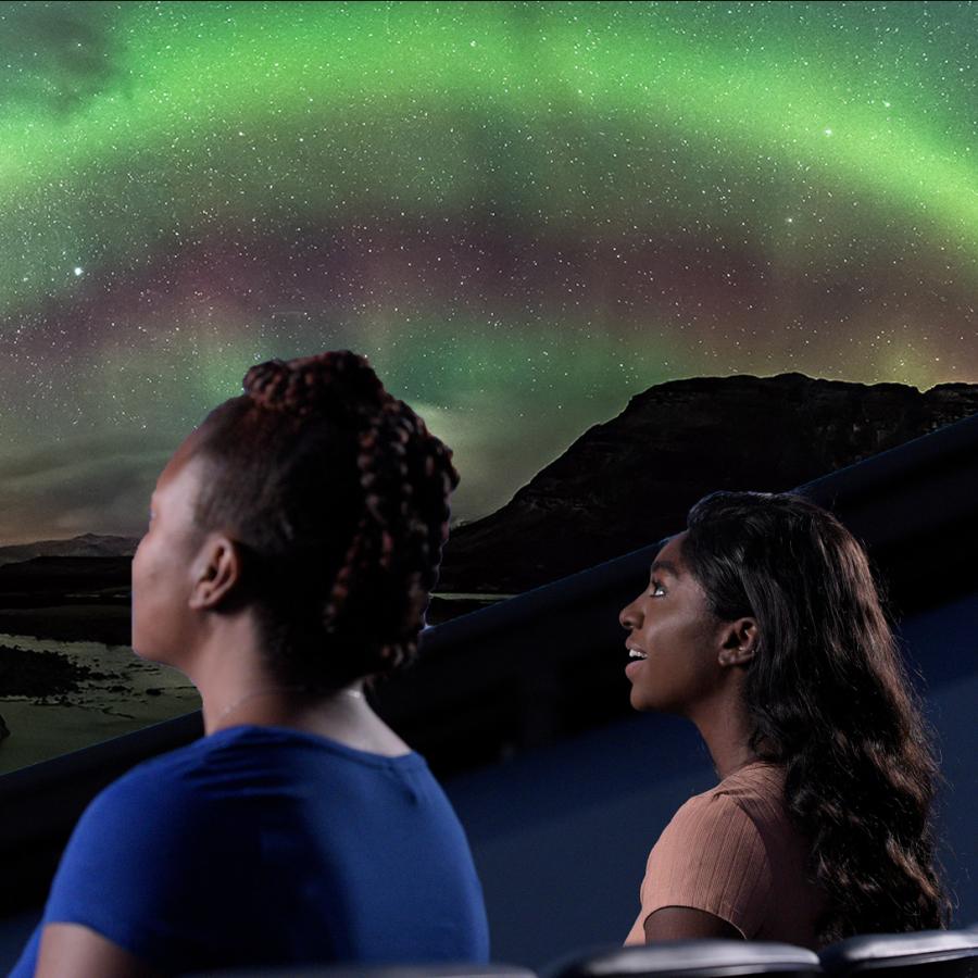 Three audience members sit in a row from left to right looking up at a screen showing a projection of bright glowing green lights