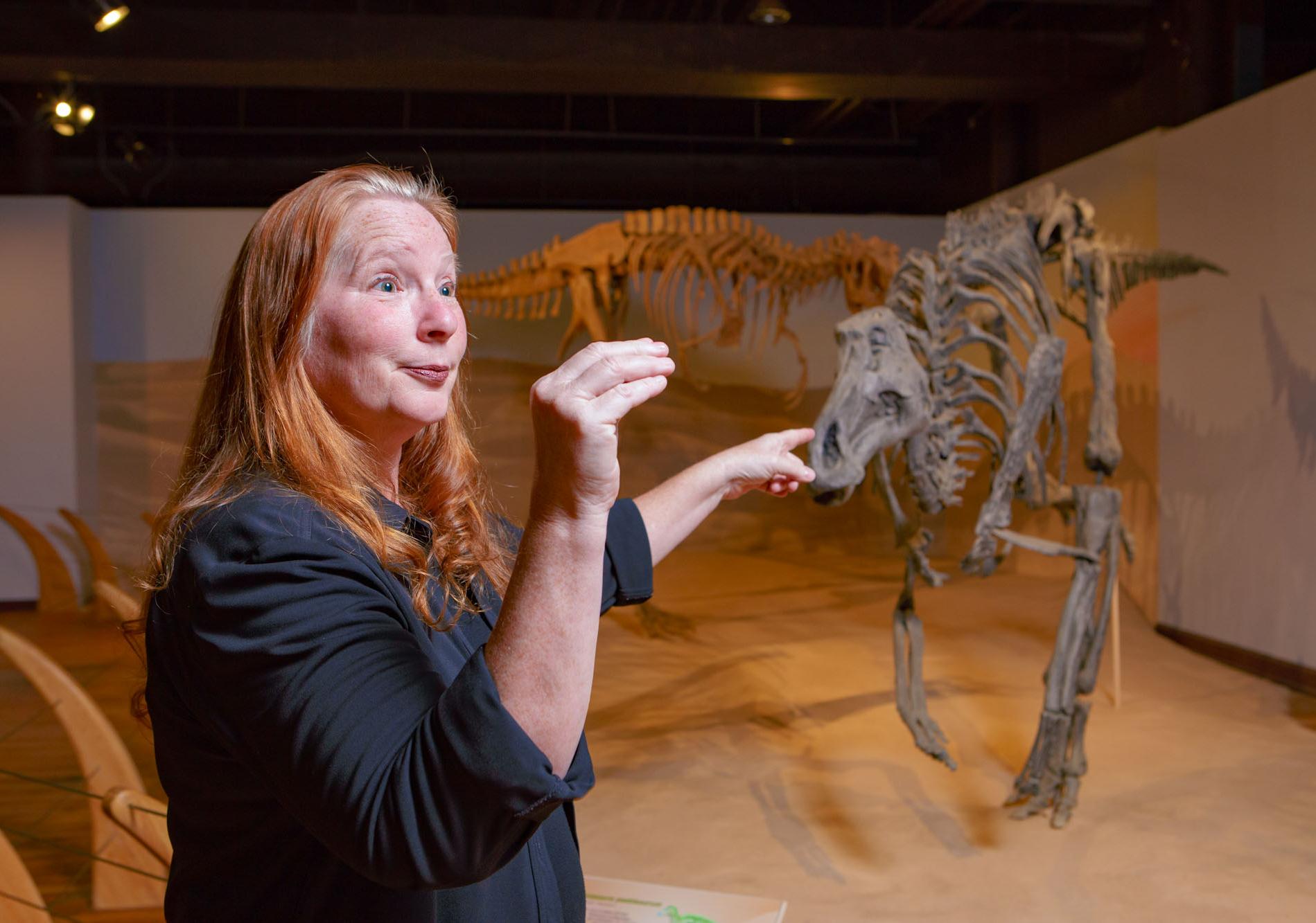 Woman stands in front of display of replica life-size dinosaur fossil skeletons with her arm and hand making a upside down L shape
