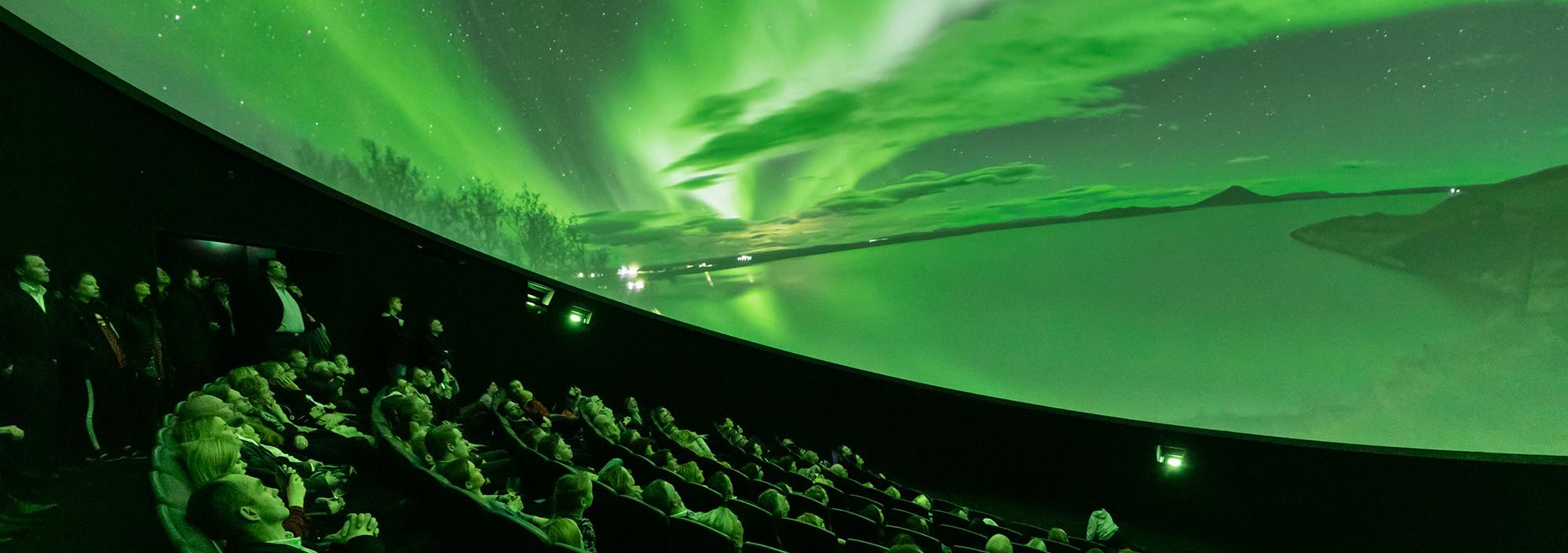 Audience looks up at domed shaped image depicting bright green lights in the sky over a snowy landscape