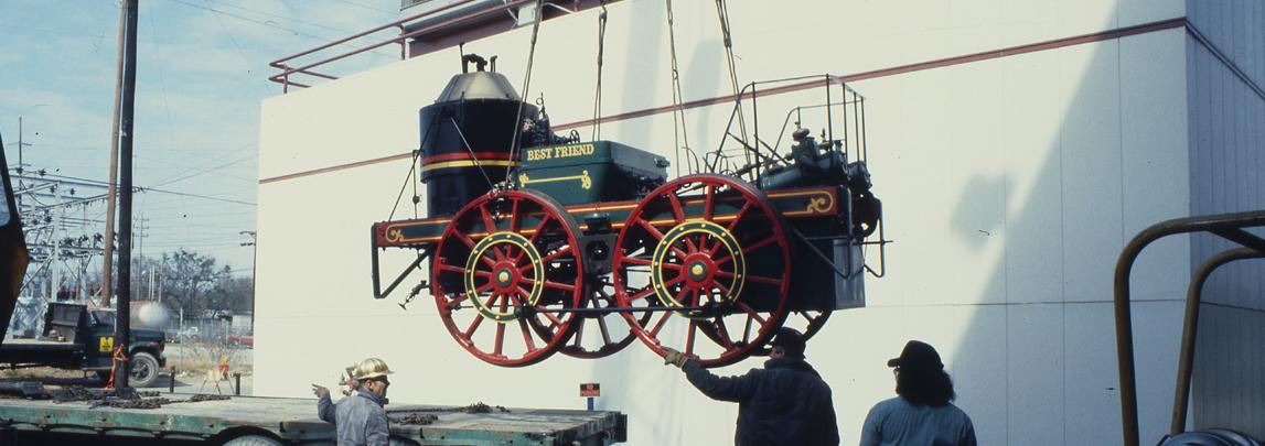 Life size antique train engine car is suspended on a lift as it is moved from the back of a flatbed truck to the ground. 