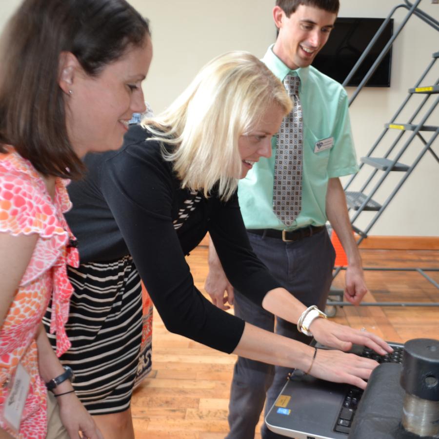 Two women look a computer screen while one types with man standing next to them in a green shirt and tie.