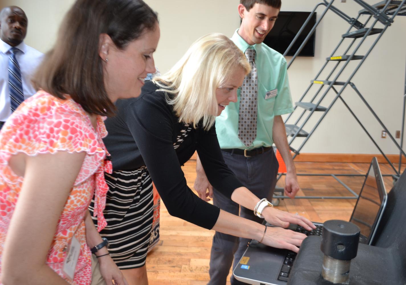 Two women look a computer screen while one types with man standing next to them in a green shirt and tie.