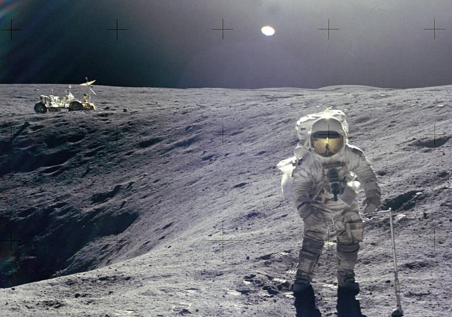 Astronaut Charles Duke stands on the Moon facing the camera next to a crater with the lunar rover at a distance in the background.