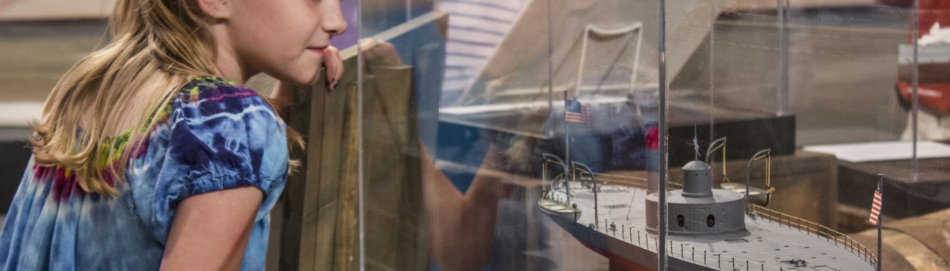 Young. blond girl in blue shirt looks into display case with model of ship inside