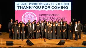 Large group of adults with women wearing black dresses and the men in tuxedos standing on a stage with a large screen behind them that reads Thank You for Coming