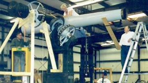 Large white telescope on heavy metal pier with several men on ladders and standing below work on it.