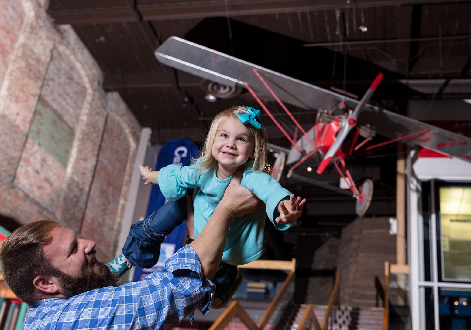 Father holds small daughter aloft as she spreads her arms like wings with the Clemson Airplane visible suspended from the ceiling behind her