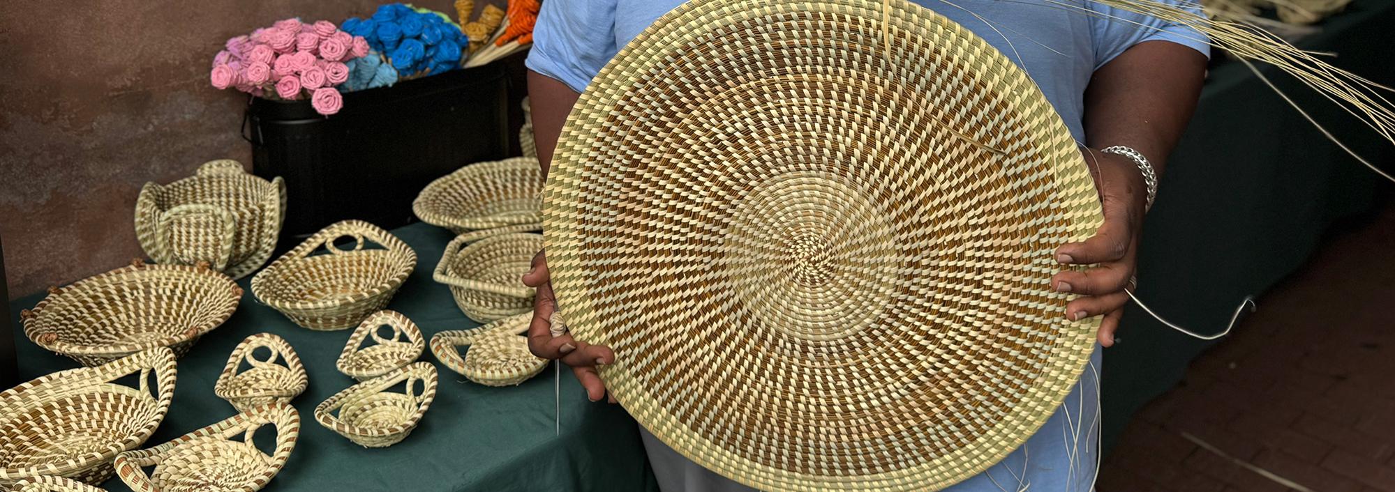 Man in blue t-shirt hold a large woven circle while standing in front of shelves displaying a variety of woven baskets, both large and small