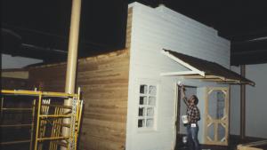 Man in plaid shirt and jeans reaches up with a paint brush outside of a partially built wooden facade for a reproduction general store