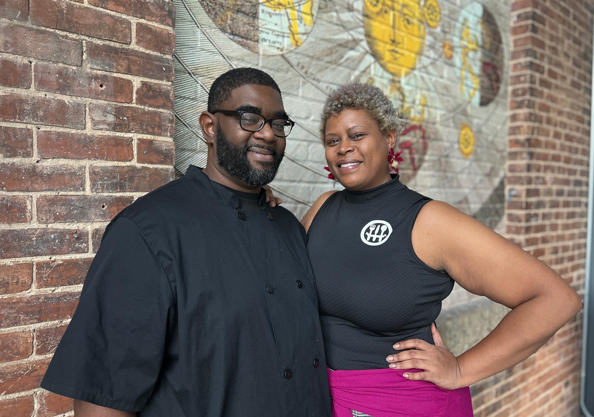 Man with glasses and woman with short gray hair pose in front of a brick wall with a design of the Sun on it