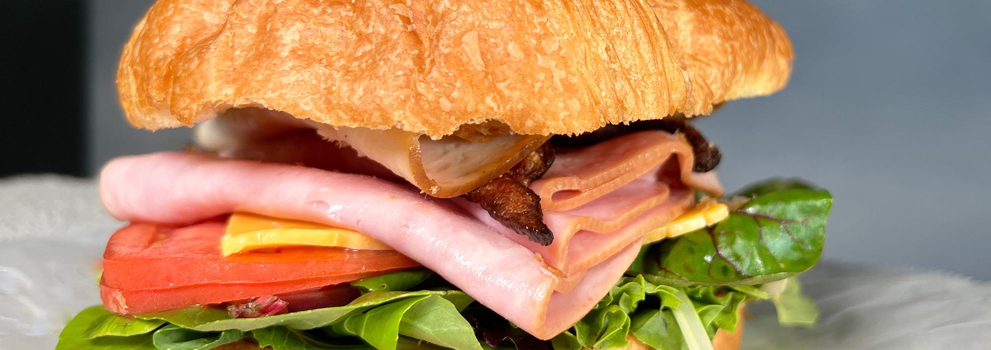 Croissant Sandwich with lettuce, tomato, ham, bacon and cheese sitting on wax paper