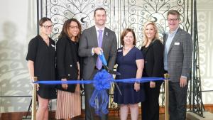 Group stands in front of a wrought iron gate on display with a blue ribbon in front of them. Man in center holds a giant pair of scissors as he prepares to cut the ribbon.