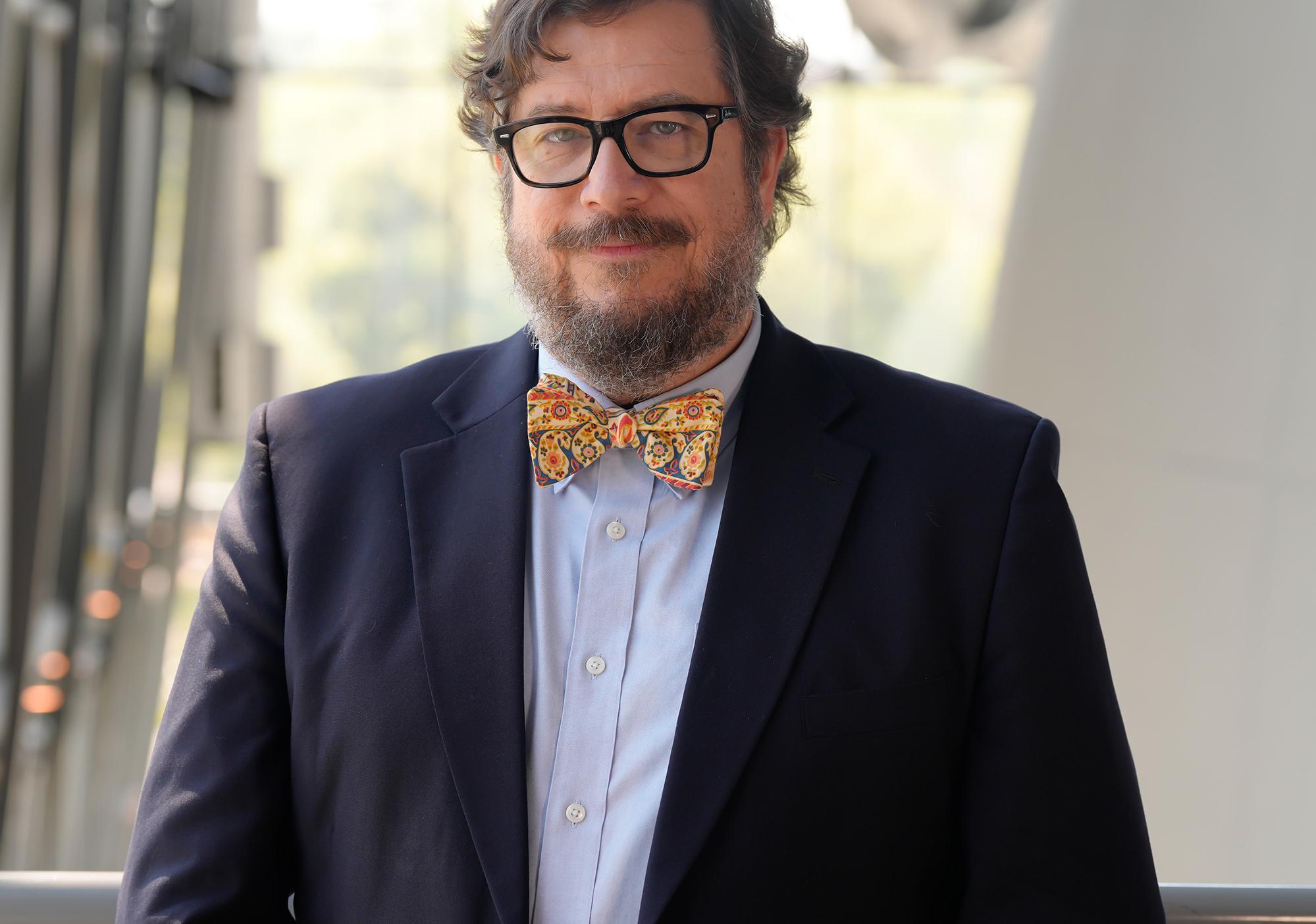 Man with beard in navy jacket and yellow bow tie smiles at camera