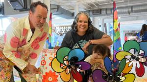 Man in a white lab coat splattered with colorful paint leans over to pose with a short, dark haired woman and a small girl in wheelchair decorated with colorful, spinning whirligigs.