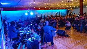 Large room lit by string lights and blue spotlights shows drummer and two guitarists performing for seated audience