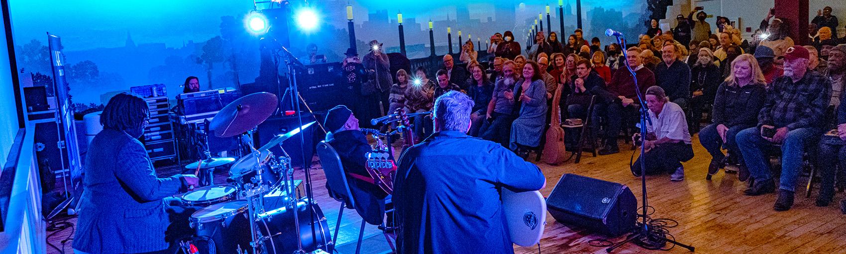 Large room lit by string lights and blue spotlights shows drummer and two guitarists performing for seated audience
