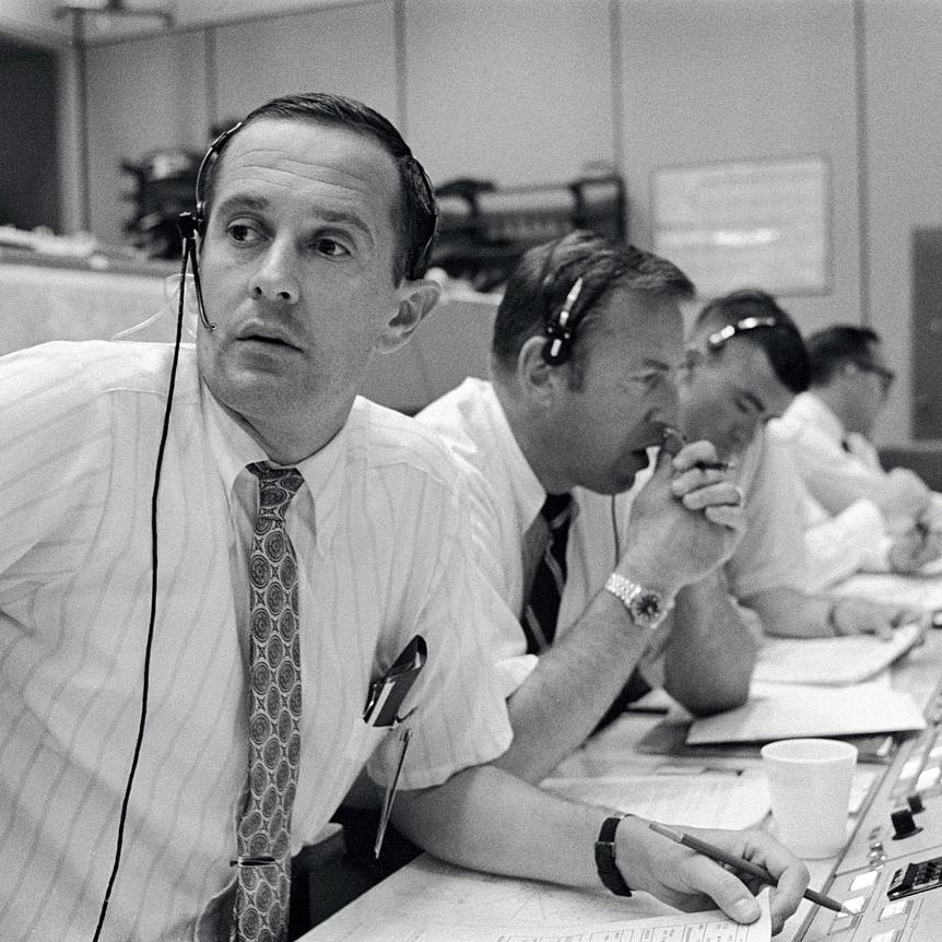 A black and white photo of four men sitting in front of control panels with headsets on