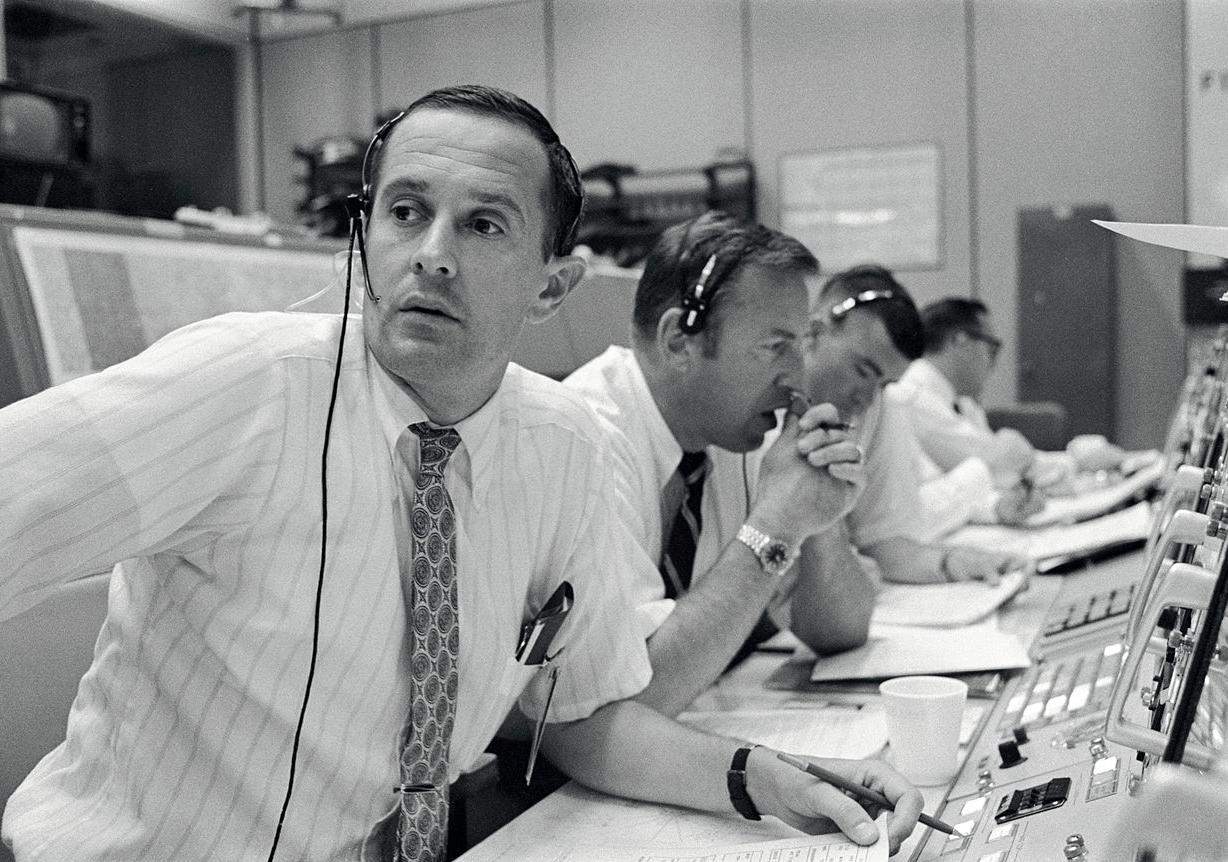 A black and white photo of four men sitting in front of control panels with headsets on