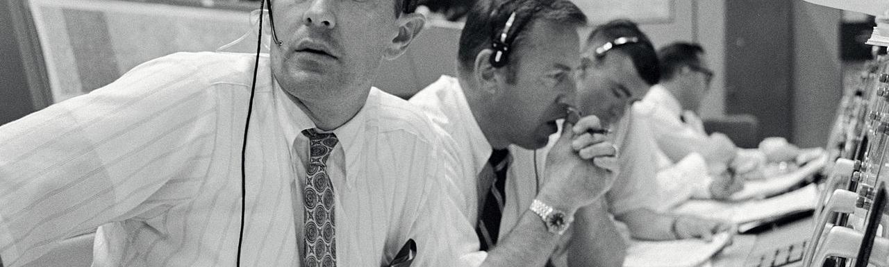 A black and white photo of four men sitting in front of control panels with headsets on