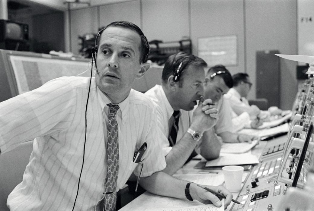 A black and white photo of four men sitting in front of control panels with headsets on