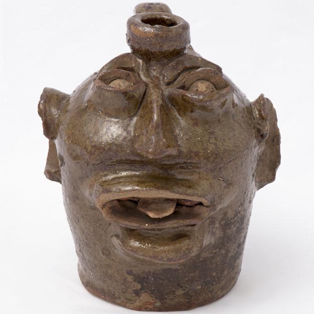 Small ceramic jug with odd looking face featuring large ears and nose, mouth open slightly with a protruding tongue.