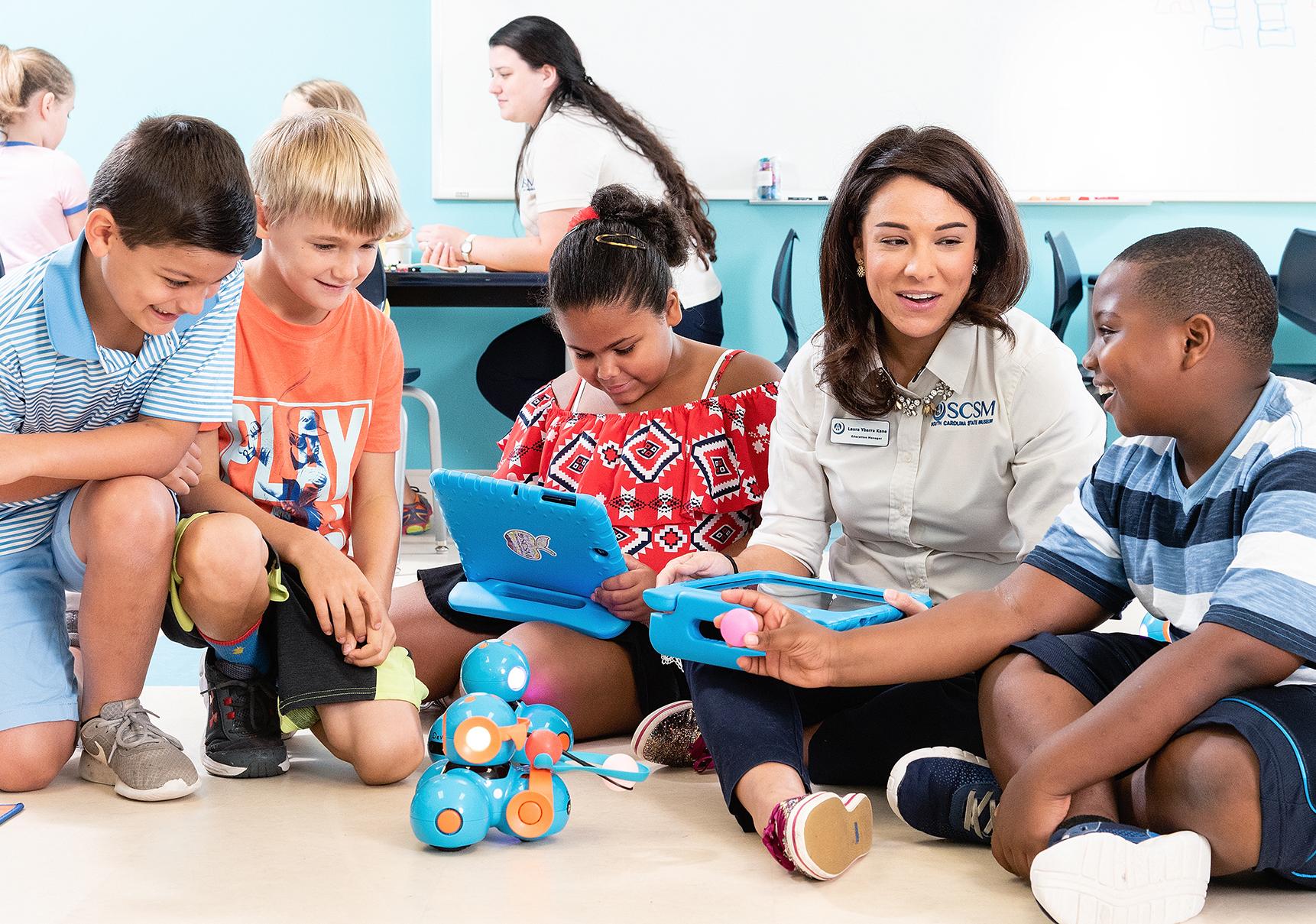 Four kids sit beside an SCSM employee viewing a tablet controlled robot