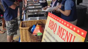 Man in tan bucket hat and blue shirt waits in a line while two older women in blue vests stand at a table serving food